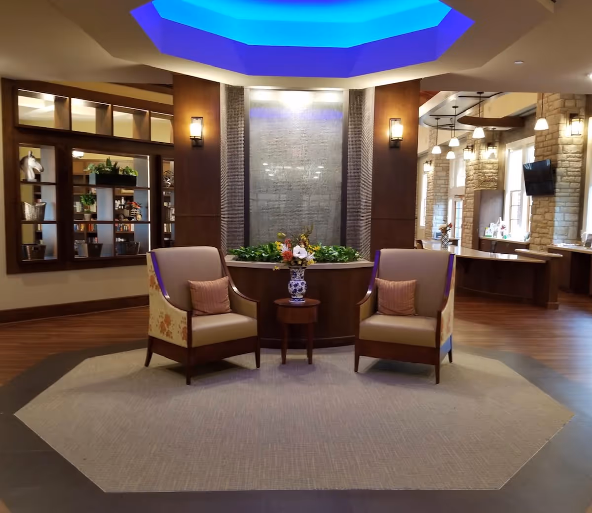 Lobby seating area with two armchairs flanking a small table and floral arrangement in front of a decorative water wall under blue ceiling lighting.