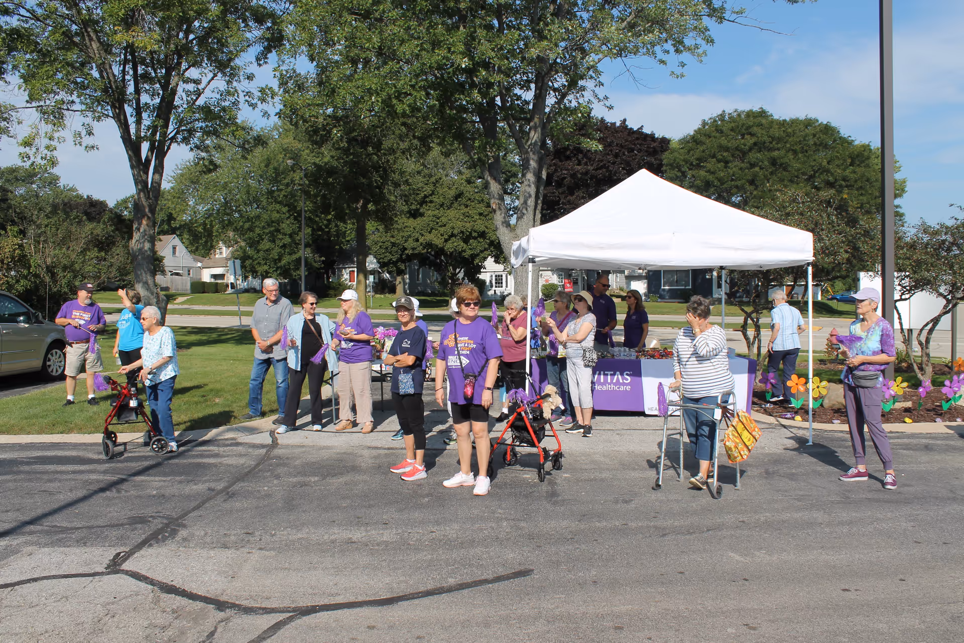 A group of seniors and volunteers gathered outdoors by a white canopy tent and table, many wearing purple shirts and using walkers.