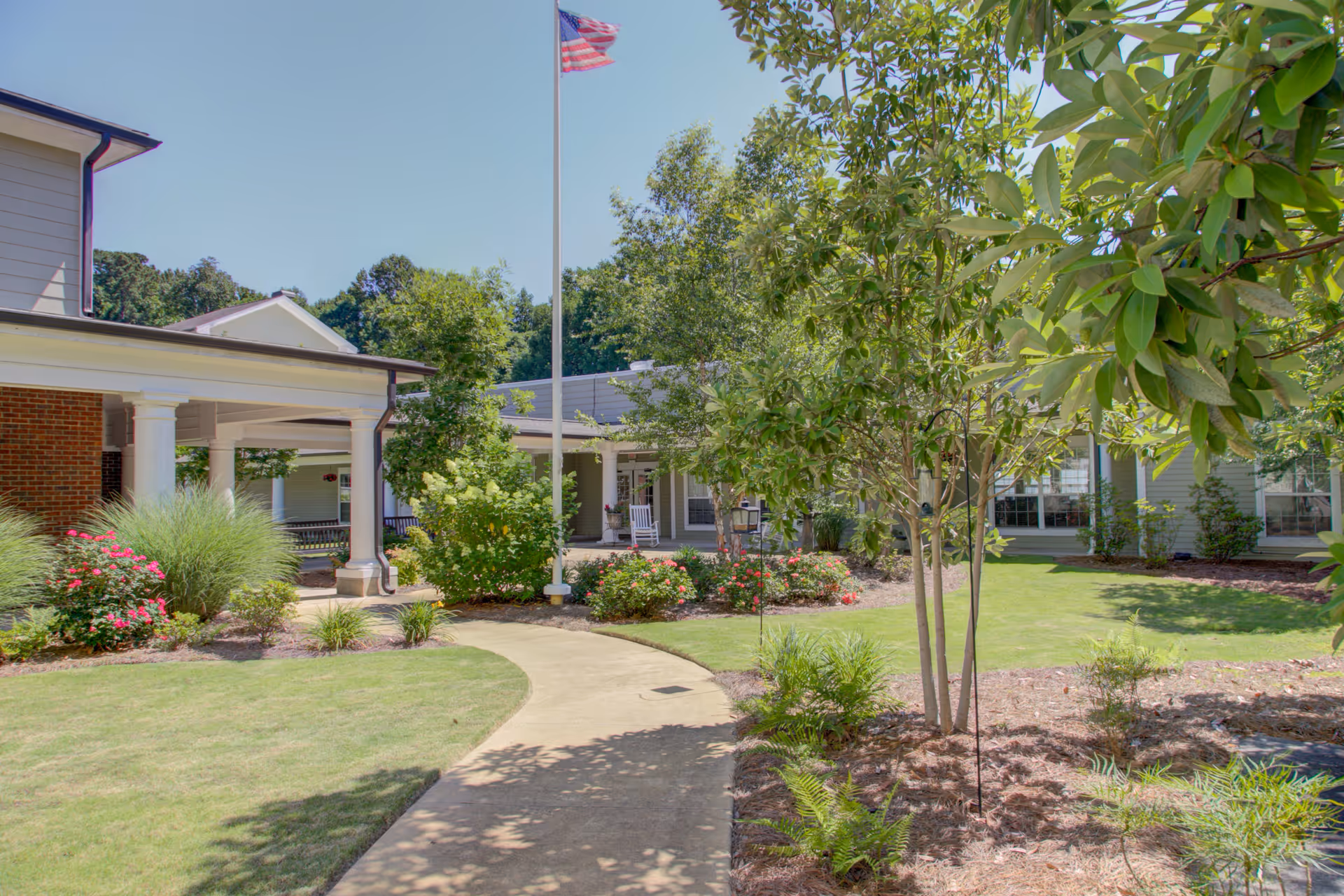Outdoor garden area at Truewood by Merrill, Riverchase with a curved concrete pathway, green grass, various shrubs and trees, an American flag on a flagpole, and part of the building with white columns and a covered porch with rocking chairs.