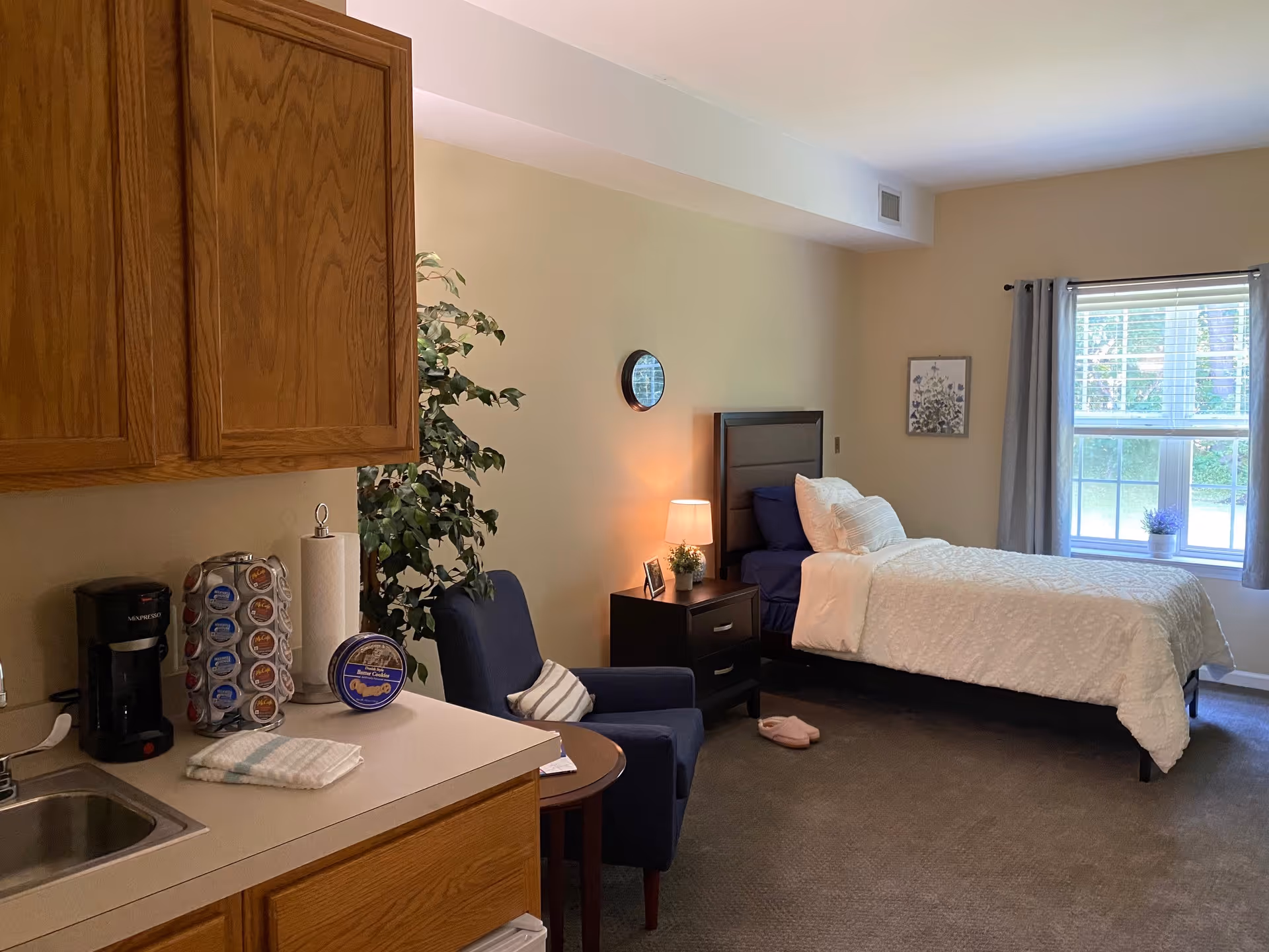 A cozy senior living bedroom featuring a single bed with white bedding and pillows, a nightstand with a lamp and small plant, a blue armchair with a striped pillow, a small round table, a window with gray curtains, and a kitchenette area with wooden cabinets, a coffee maker, a paper towel holder, and a K-cup holder.