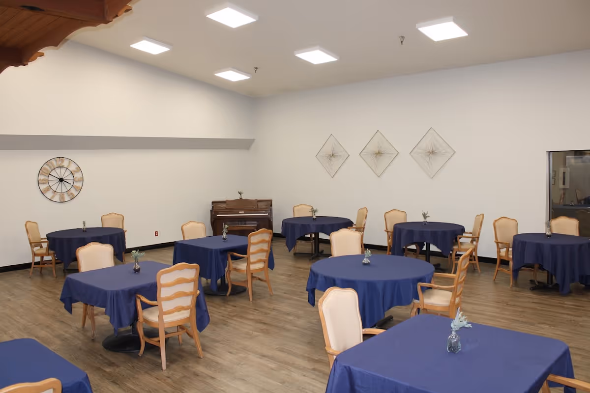 A spacious dining room with several round and square tables covered with blue tablecloths. Each table has a small vase with greenery. The room has wooden chairs with light-colored cushions, wood flooring, white walls, and ceiling lights. There is a piano against the far wall and decorative wall art including a large clock and three diamond-shaped wire sculptures.