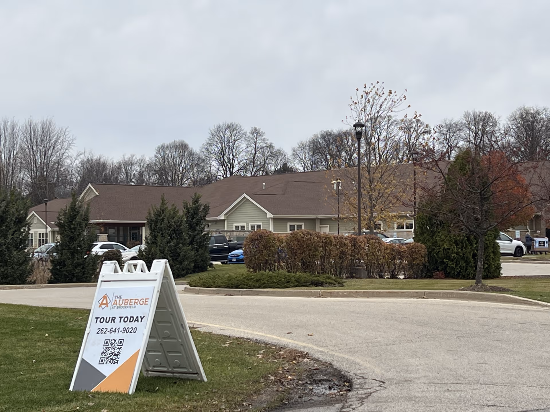 Exterior view of The Auberge at Brookfield senior living facility on a cloudy day, showing a single-story building with a brown roof, surrounded by trees and bushes. In the foreground, there is a white sandwich board sign advertising tours with a phone number and QR code.