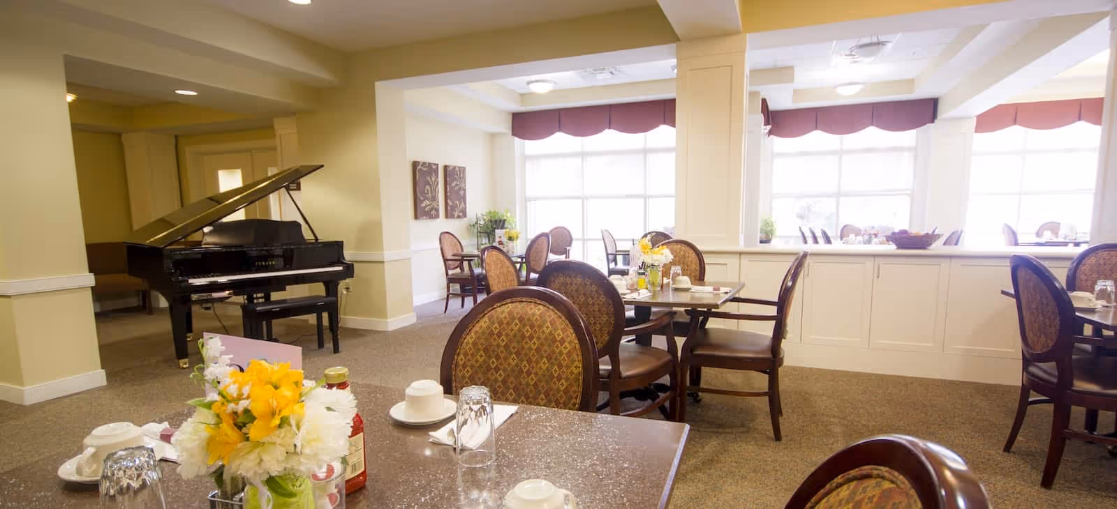 A bright dining room with multiple tables and chairs arranged neatly. Each table has place settings including cups, glasses, and napkins. A vase with yellow and white flowers is on the nearest table. In the background, there is a black grand piano and large windows with red valances letting in natural light.