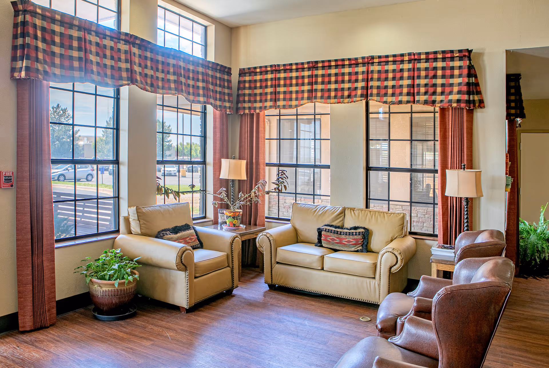 A cozy sitting area in a senior living facility with large windows covered by plaid valances and red curtains. The room features a beige loveseat and armchair with decorative pillows, two brown leather chairs, a wooden side table with a lamp, and potted plants on the floor and table. The floor is wooden, and natural light fills the space.