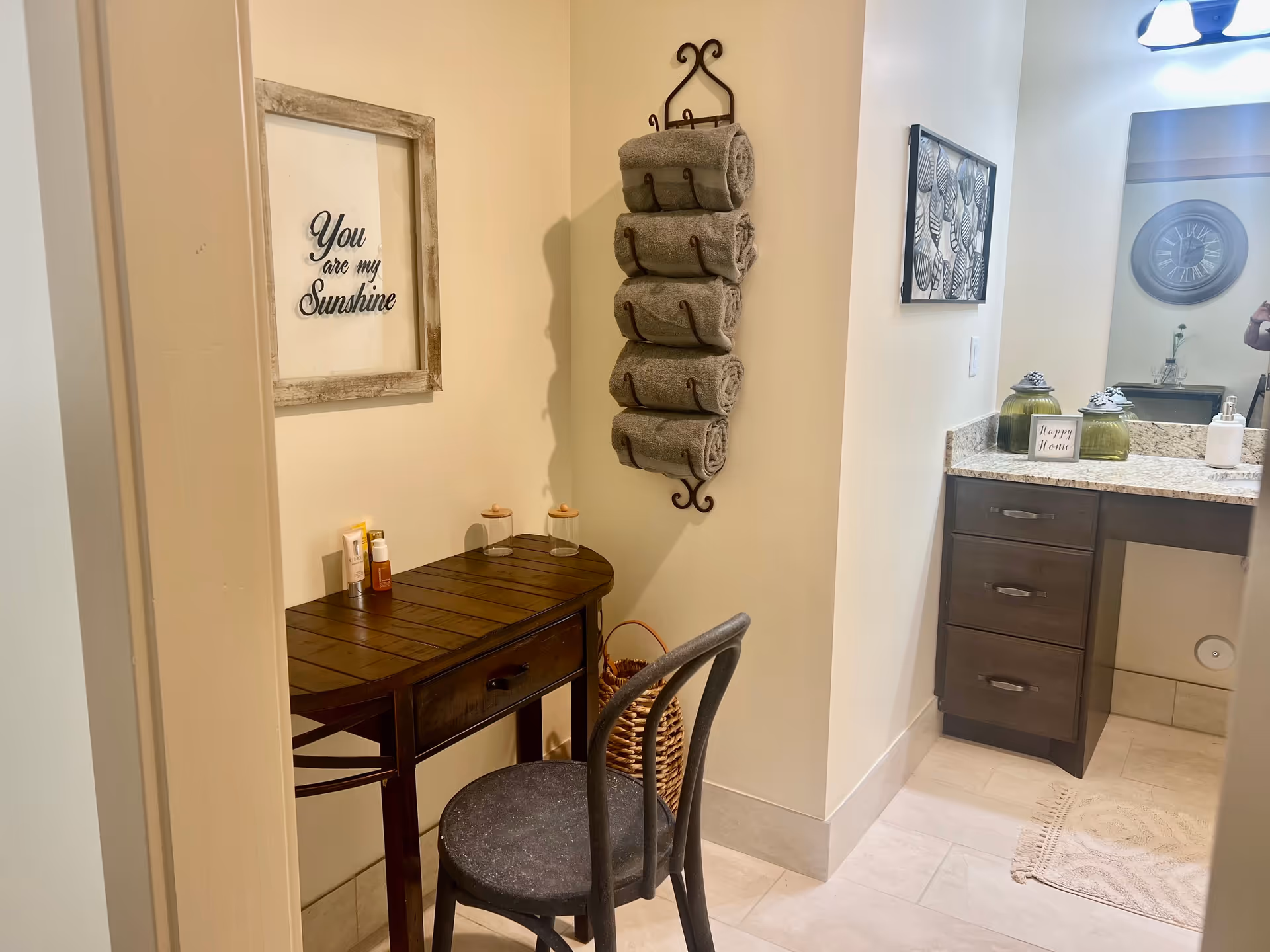 A cozy bathroom corner with a small dark wooden table and chair. On the wall above the table is a framed sign that reads 'You are my Sunshine.' Next to the table, a decorative wall-mounted rack holds five rolled gray towels. To the right is a bathroom vanity with a granite countertop, dark wood drawers, a mirror, and a small sign that says 'Happy Home.' The floor is tiled and a beige rug is placed in front of the vanity.