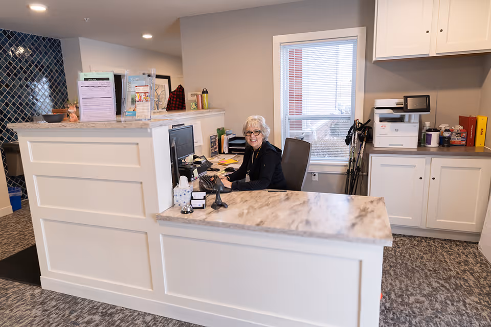 A smiling woman with gray hair and glasses sits behind a white reception desk with a marble countertop in a well-lit office area. The desk has a computer, a tissue box, and various office supplies. Behind her, there is a window with blinds, white cabinets, a printer, and some walking sticks. The floor is carpeted with a patterned design.