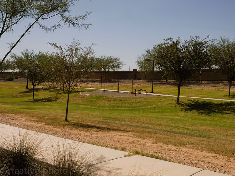Grassy outdoor area with trees, a walking path and small playground or exercise equipment under a clear sky.