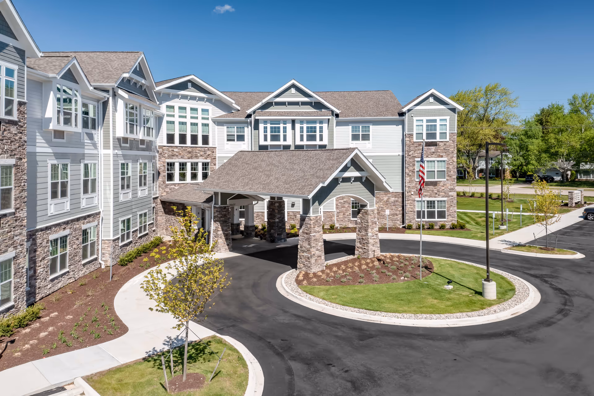 Front entrance and porte-cochère of a multi-story senior living building with stone accents, circular driveway, landscaping, and an American flag.