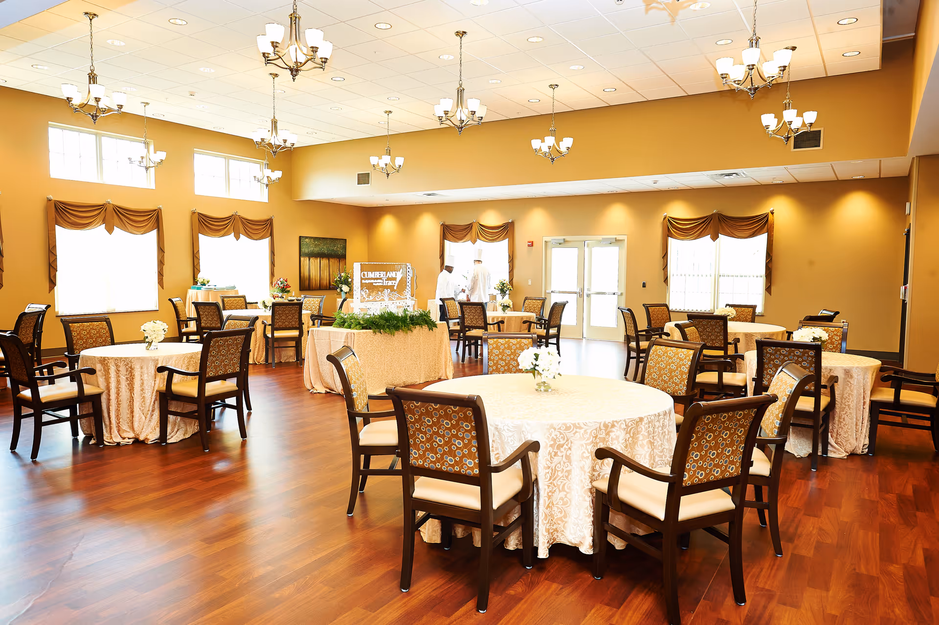 Bright senior living dining/event room with round tables covered in tablecloths and wooden chairs under chandeliers.