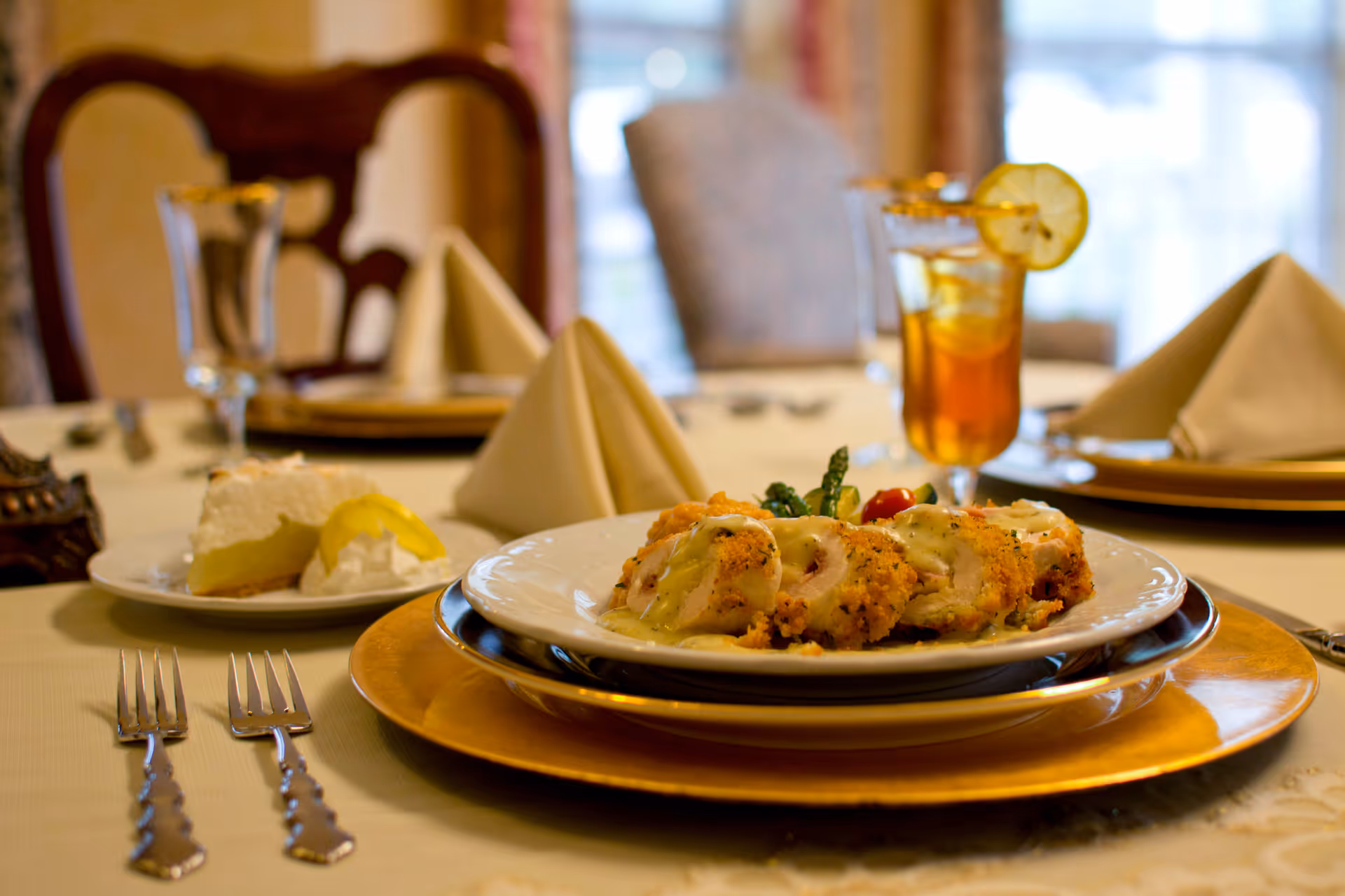 A close-up view of a dining table set with a plated meal consisting of breaded chicken rolls with sauce and vegetables, a slice of lemon meringue pie with whipped cream, two forks, a glass of iced tea garnished with a lemon slice, and neatly folded beige napkins on gold-rimmed plates in an elegant dining room setting.