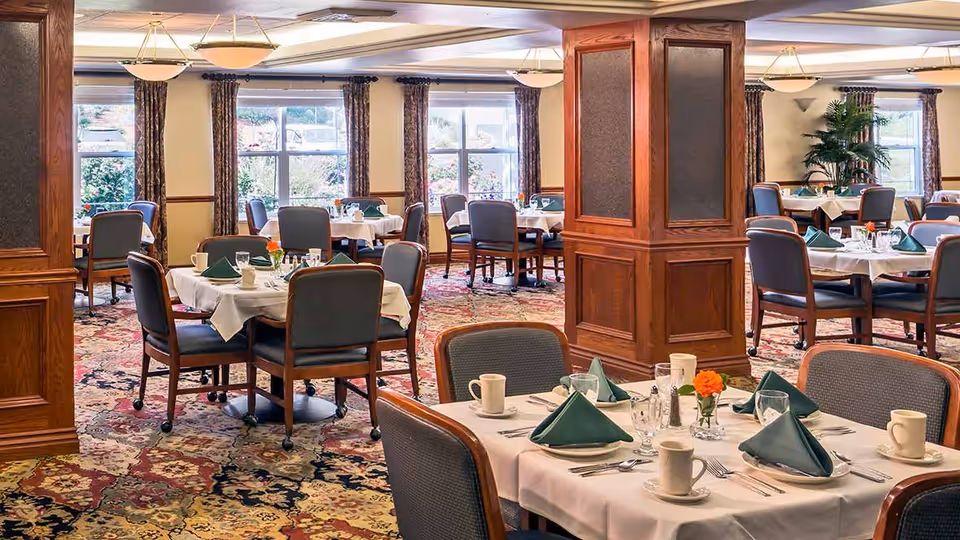 A spacious dining room with multiple tables set for a meal. Each table is covered with a white tablecloth, green folded napkins, cups, glasses, and silverware. The room features large windows with patterned curtains, wooden pillars, and a colorful patterned carpet. There are chairs around each table, and a potted plant is visible near one window.