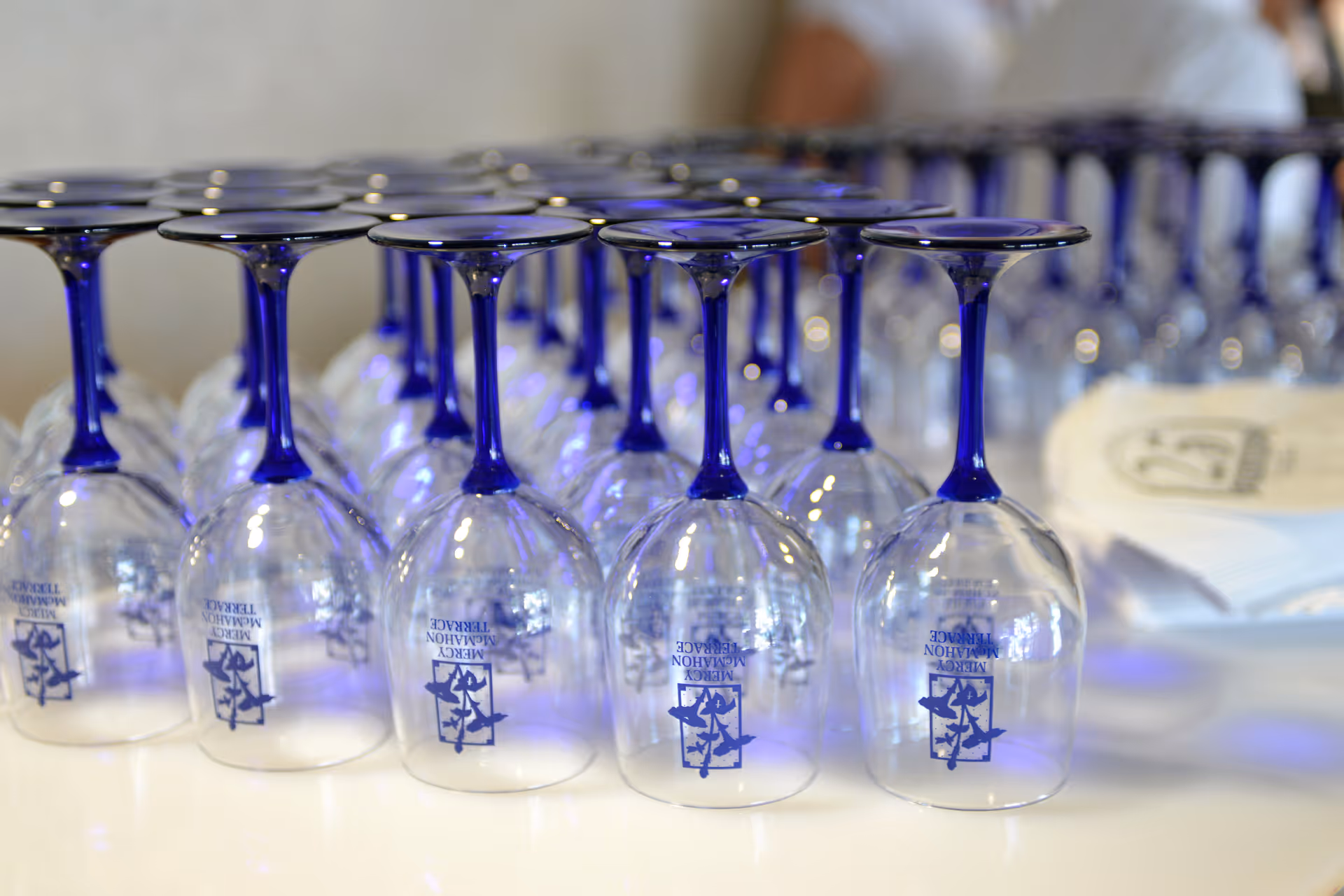 Rows of upside-down wine glasses with blue stems neatly arranged on a white surface, each glass labeled with 'Mercy McMahon Terrace'.
