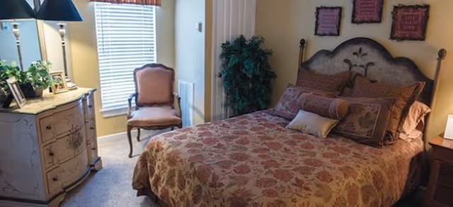 Bedroom featuring a floral-patterned bed with decorative pillows and headboard, a dresser and lamp, an upholstered chair by a window, and wall decor.