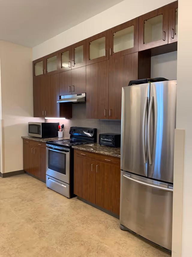 A modern kitchen with wooden cabinets, a stainless steel refrigerator, an electric stove with oven, a microwave, and a toaster oven on the countertop. The floor is tiled and the walls are light-colored.