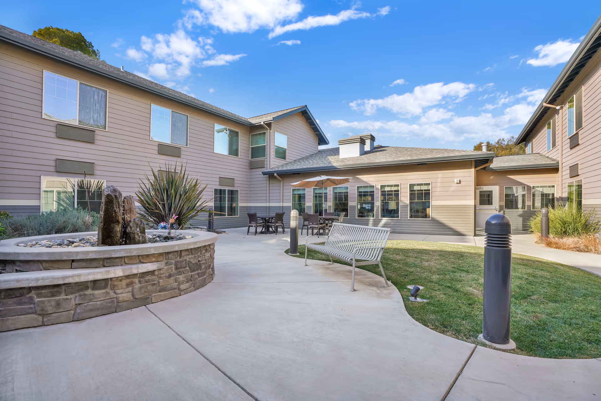 Outdoor courtyard area of a senior living facility with a stone water fountain, metal bench, patio tables with umbrellas, and a two-story building surrounding the courtyard under a blue sky with scattered clouds.