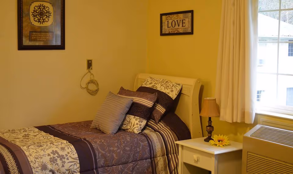 Small furnished bedroom with a single bed covered in patterned bedding, a nightstand with a lamp and a window with curtains.
