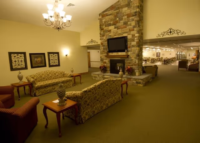 A spacious assisted-living common room with floral sofas and chairs arranged around a stone fireplace topped by a wall-mounted TV, with a dining area visible in the background.