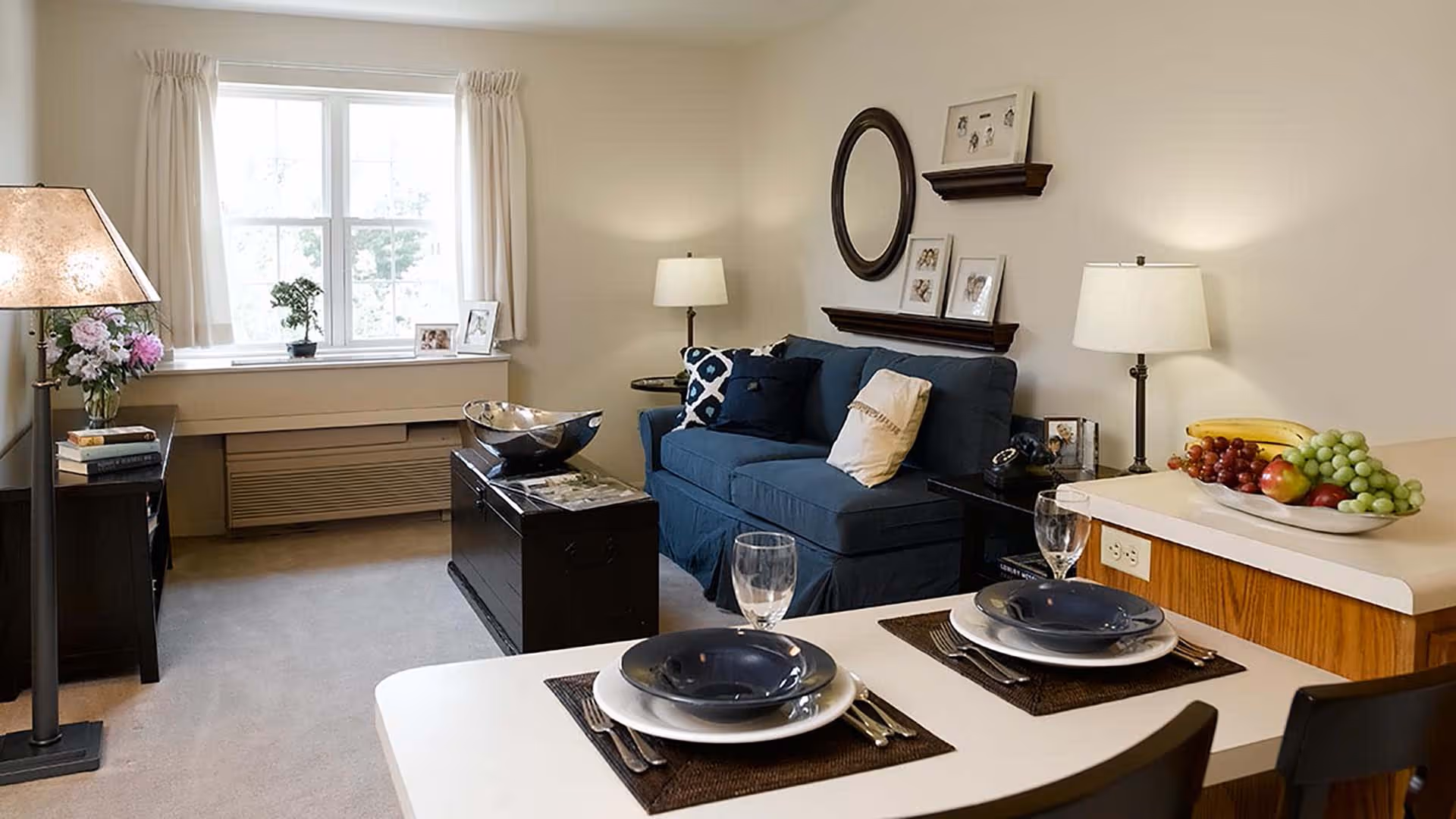 A cozy living room with a blue sofa adorned with pillows, a black coffee table, and a side table with a lamp. There is a window with cream curtains letting in natural light. In the foreground, a dining counter is set with two place settings including plates, bowls, glasses, and cutlery. A bowl of fruit is placed on the counter. The walls have decorative shelves with framed pictures and a round mirror.