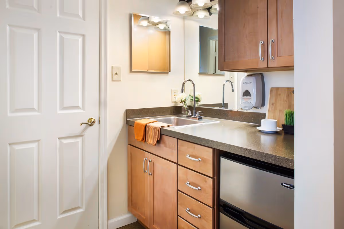 A small kitchenette with a sink, dark countertop, wooden cabinets, a wall-mounted soap dispenser and a compact stainless steel refrigerator.