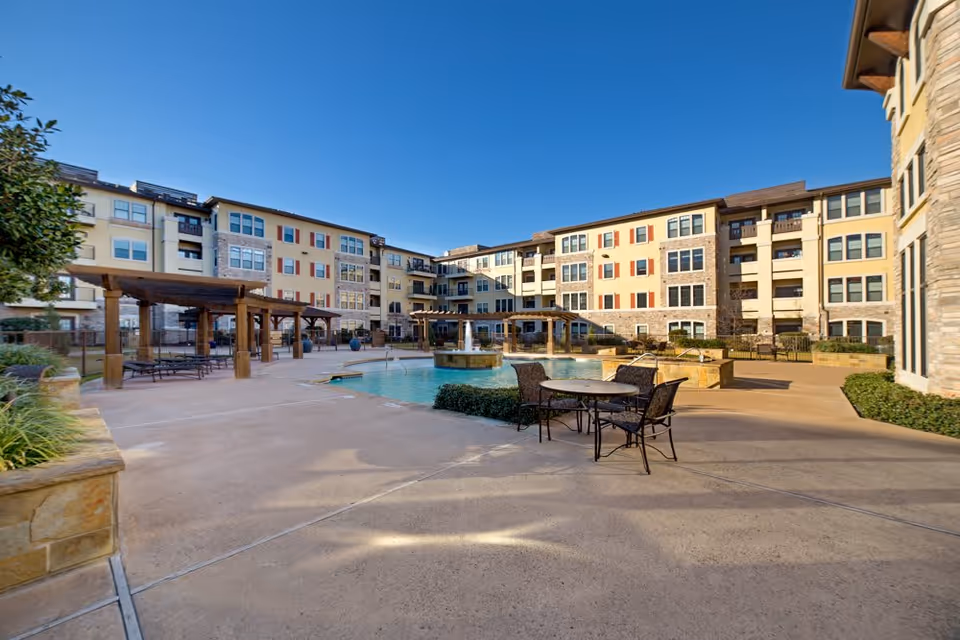 Outdoor courtyard area of a senior living facility with a swimming pool, patio tables with chairs, pergolas, and a multi-story building in the background under a clear blue sky.