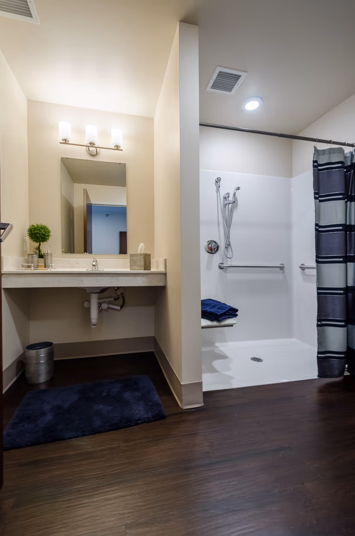 A clean and modern bathroom featuring a vanity with a sink, a mirror with three light fixtures above it, a small potted plant, soap dispenser, and tissue box on the countertop. To the right is a walk-in shower with a handheld showerhead, grab bars, a fold-down seat with folded towels, and a striped shower curtain. The floor is dark wood, and there is a dark blue bath mat in front of the vanity.