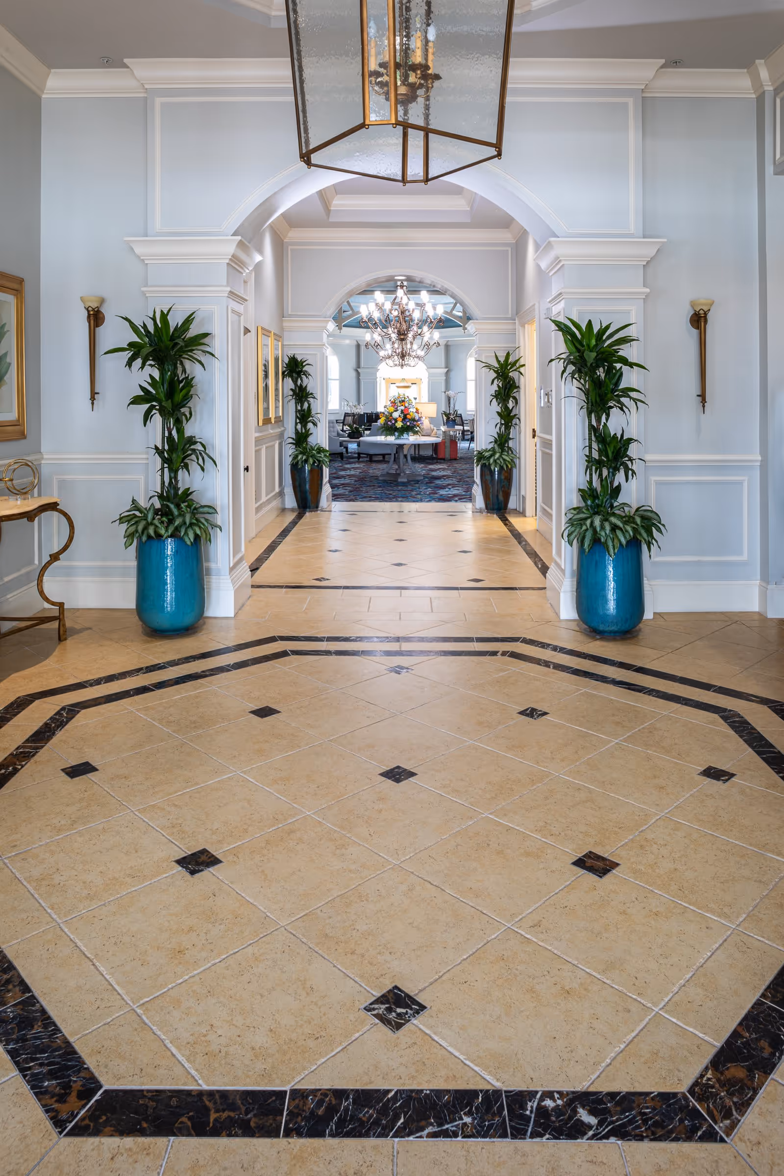 Elegant interior hallway with beige tiled floor bordered by dark marble tiles, light blue walls with white trim, two large blue planters with green plants on either side of an archway leading to a room with a chandelier and a round table with a floral arrangement.