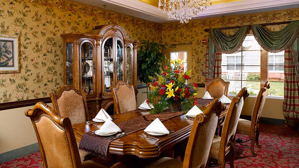A formal dining room with a large wooden dining table set for six people with folded white napkins and silverware. The room features ornate wooden chairs with upholstered seats, a floral centerpiece on the table, a wooden china cabinet displaying plates, floral wallpaper, a chandelier, and large windows with green and red draped curtains.