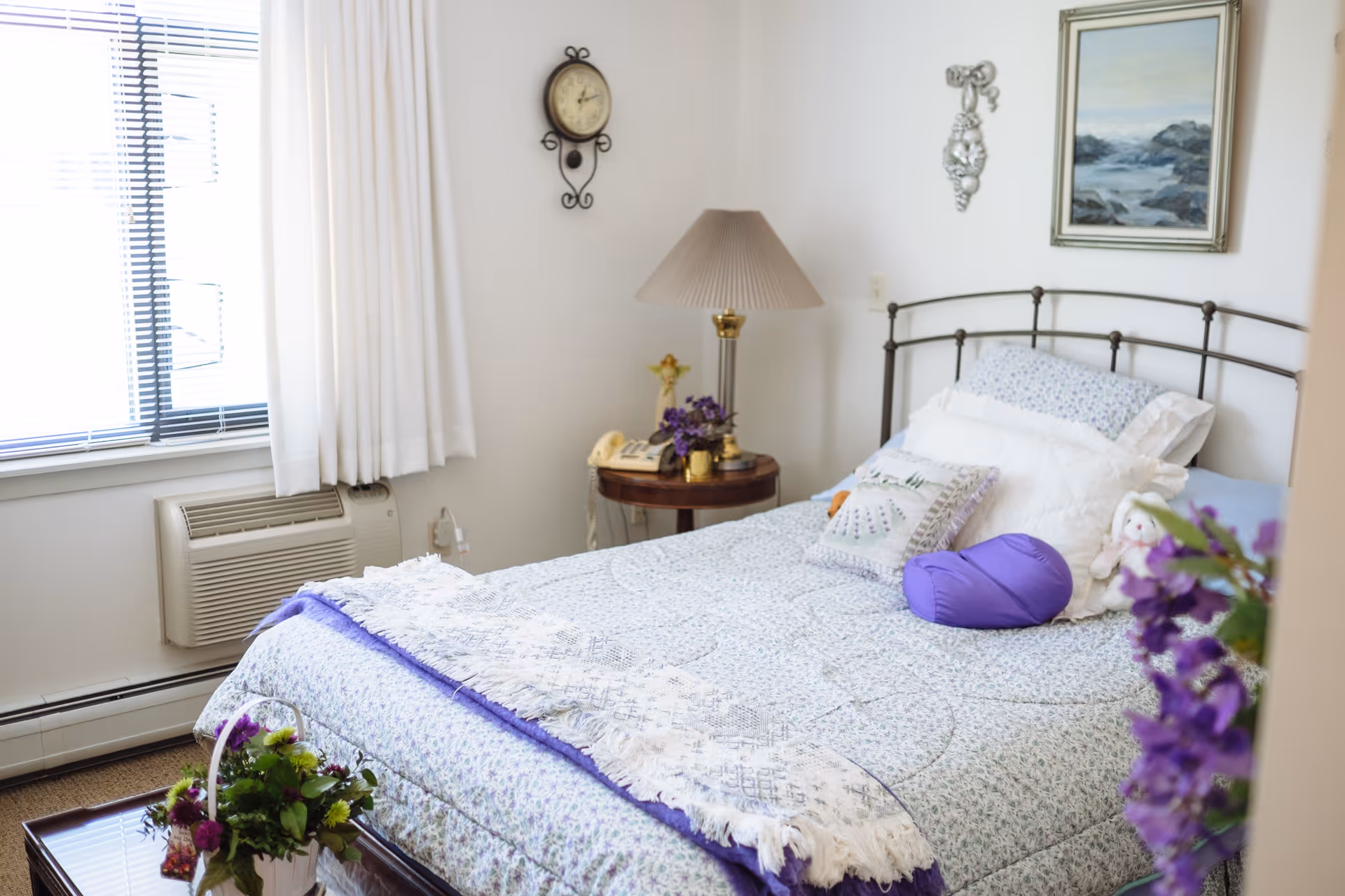 A cozy bedroom with a neatly made bed featuring a floral patterned bedspread and multiple pillows, including a purple cylindrical pillow. There is a wooden side table with a lamp, a telephone, and a small flower arrangement. A wall clock and framed artwork hang on the white walls. A window with white curtains and blinds lets in natural light, and a small air conditioning unit is installed below the window. A vase with purple flowers is partially visible in the foreground.