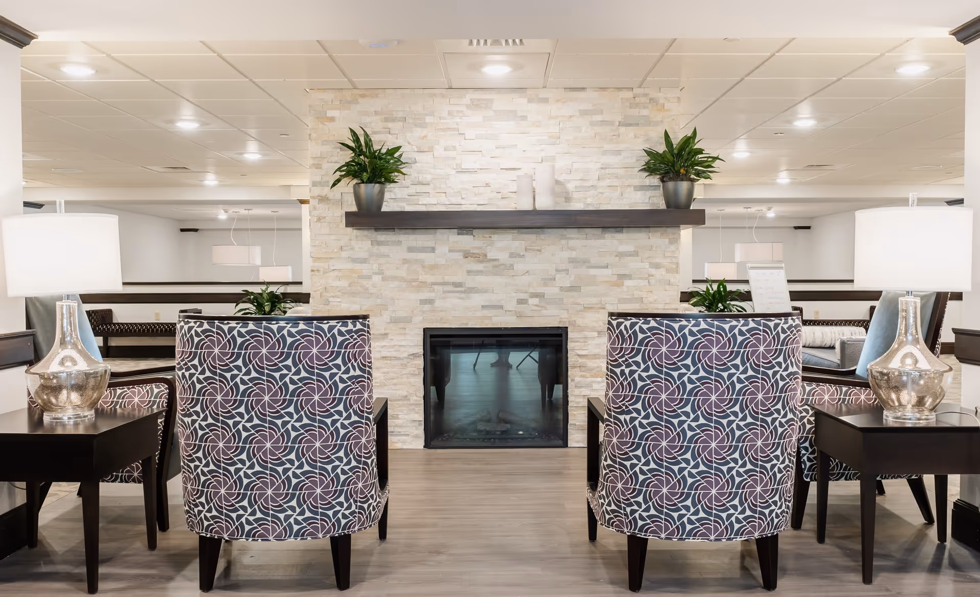A cozy indoor seating area featuring two patterned armchairs facing a modern stone fireplace with a dark wooden mantel. Each chair is accompanied by a dark wooden side table with a silver lamp. Two potted plants are placed on the mantel, and the room has a light-colored ceiling with recessed lighting.