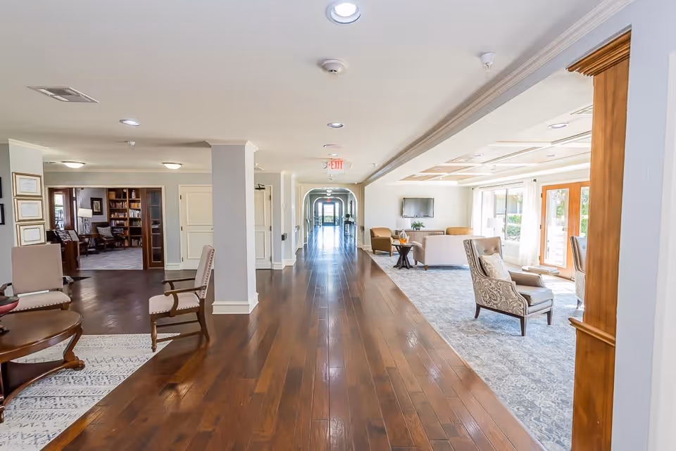 A bright and spacious interior hallway of a senior living facility with polished wooden floors and comfortable seating areas on both sides. On the left, there is a cozy room with bookshelves and chairs, while on the right, a lounge area with armchairs, a sofa, a coffee table, and a wall-mounted TV is visible. Large windows and glass doors allow natural light to fill the space.