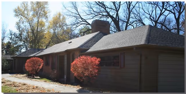 Exterior view of a single-story residential building with a sloped roof, chimney, and red bushes in front. Trees with autumn foliage are visible in the background under a clear sky.