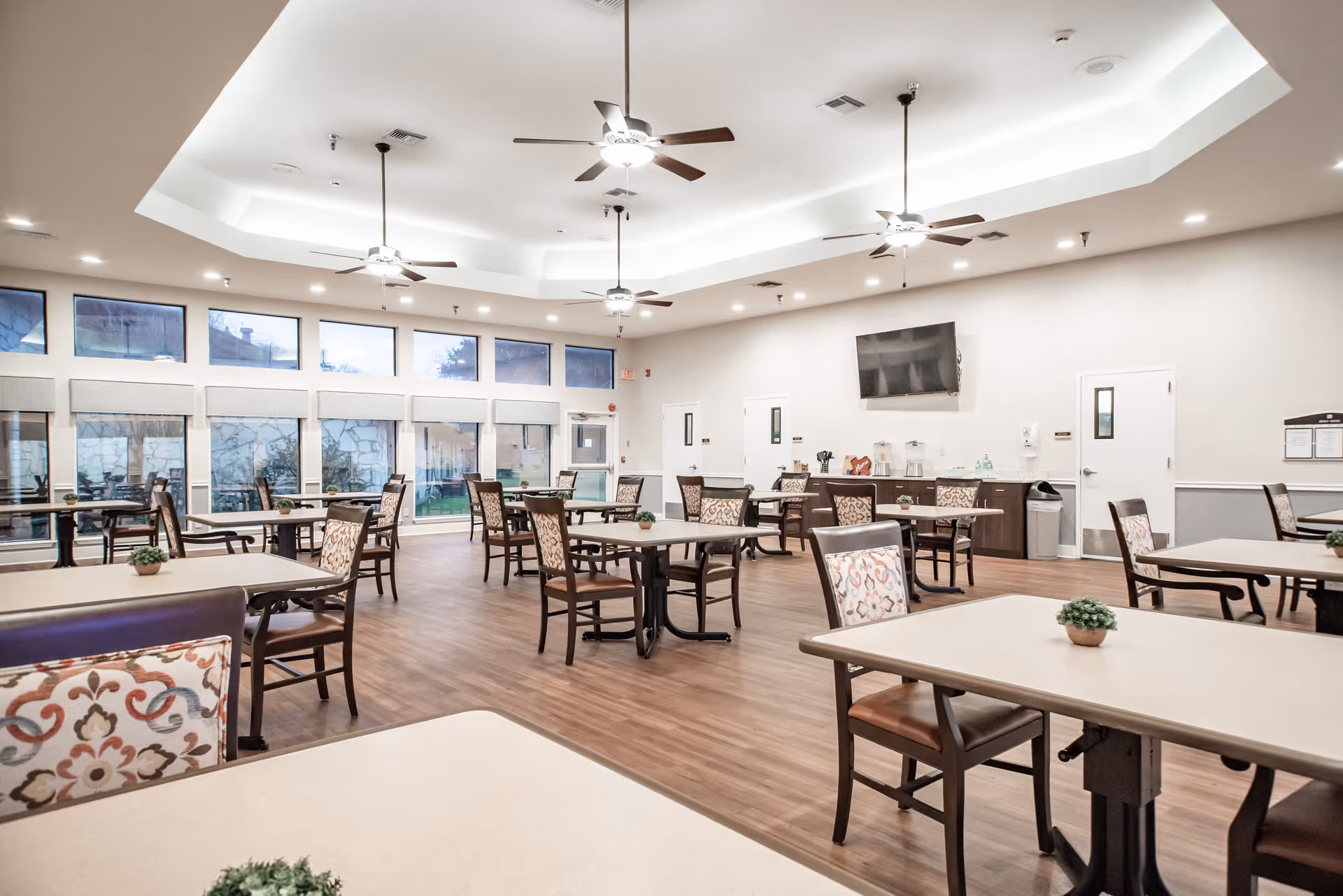 A spacious and well-lit dining room with multiple tables and chairs arranged neatly. The room features large windows along one wall, ceiling fans with lights, and a wall-mounted television. Small potted plants are placed on the tables, and the floor is wooden.