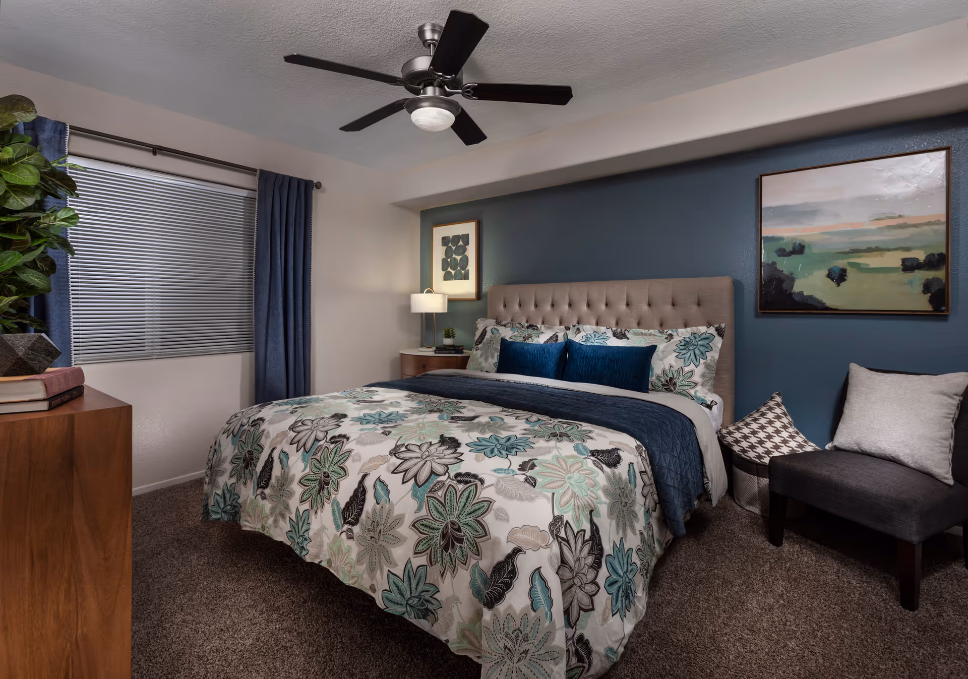 A cozy bedroom with a large bed featuring a floral patterned comforter in shades of teal, gray, and white. The bed has a tufted beige headboard and is adorned with decorative pillows in blue and floral patterns. To the right of the bed is a small gray chair with two pillows, one in a houndstooth pattern and the other in solid gray. The wall behind the bed is painted dark teal and decorated with two framed artworks. A wooden dresser with books and a plant is on the left side of the room, next to a window with closed blinds and dark blue curtains. A ceiling fan with a light fixture is mounted on the white ceiling.