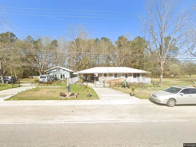 Front exterior view of Serenity Manor At Holly Hill, showing a single-story building with a white roof and a ramp leading to the entrance. There are trees and grass surrounding the building, a driveway with parked cars, and a clear blue sky above.