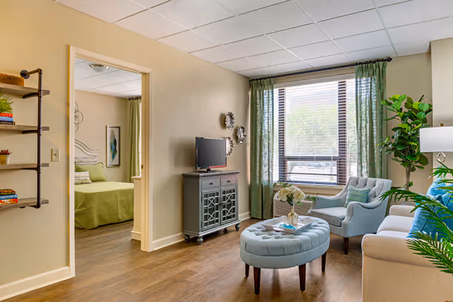 A cozy living room with a beige sofa, a light blue armchair, and a matching ottoman in the center. A small cabinet with a TV on top is against the wall, and a large window with green curtains lets in natural light. To the left, an open doorway leads to a bedroom with a green bedspread. The room has wooden flooring and light-colored walls, decorated with plants and wall art.