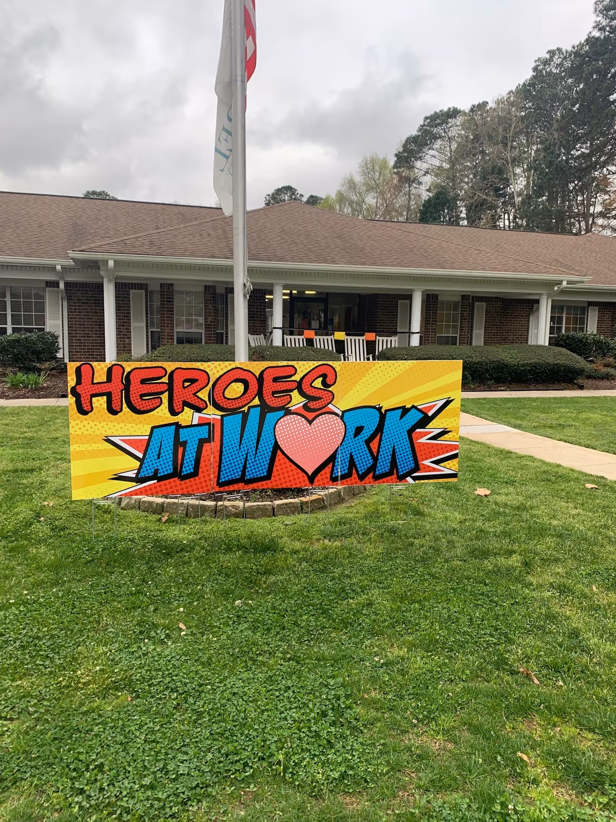 A colorful sign on a grassy lawn in front of a brick building with white columns and a brown roof. The sign reads 'HEROES AT WORK' with a heart symbol replacing the letter O in 'WORK'. There is a flagpole behind the sign with a partially visible flag, and the sky is cloudy.