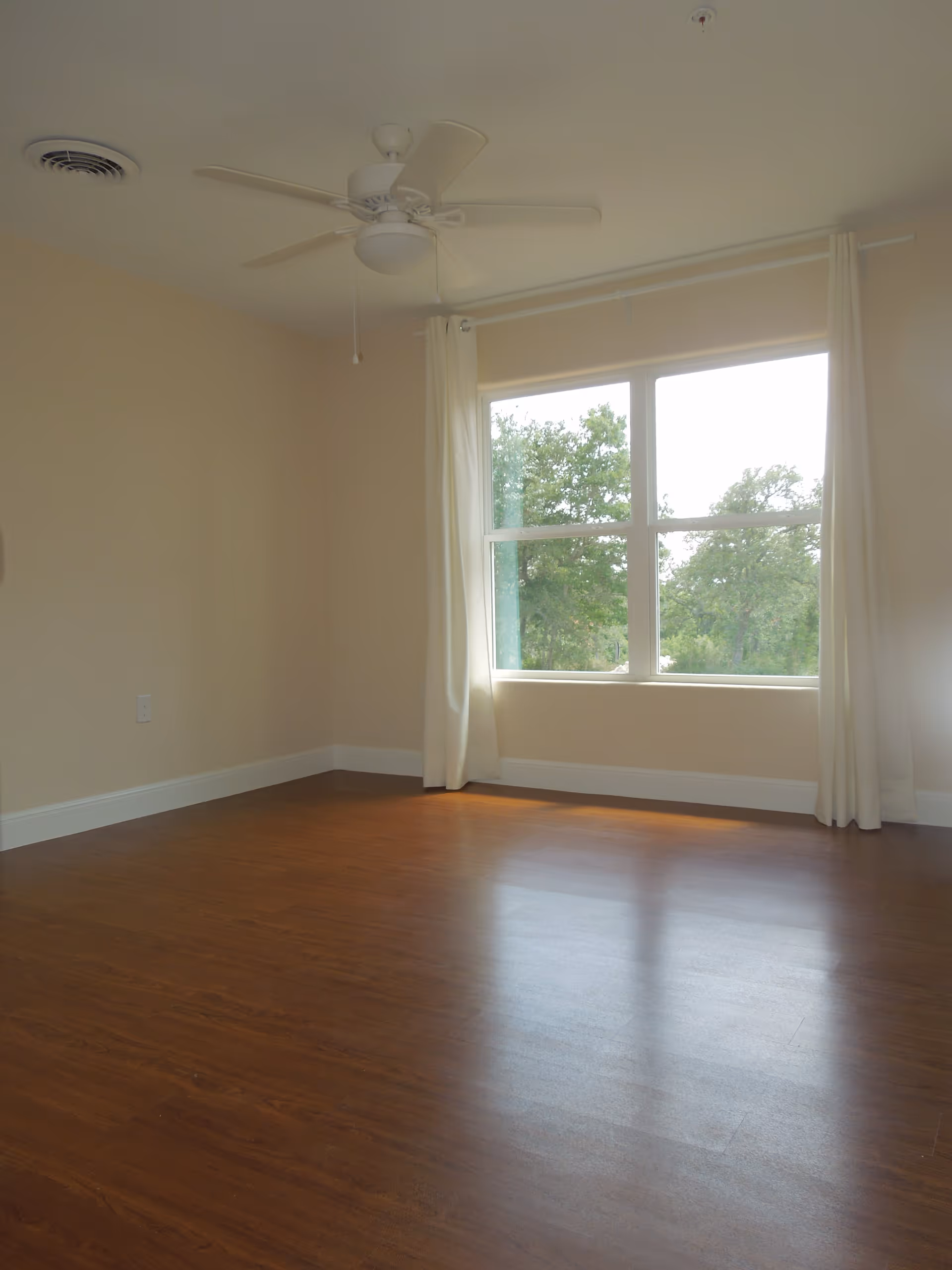 Empty room with wooden floor, beige walls, a large window with white curtains, and a white ceiling fan.