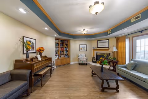 Spacious common living room with a piano, bookshelves, fireplace, seating, and a coffee table by a large window.