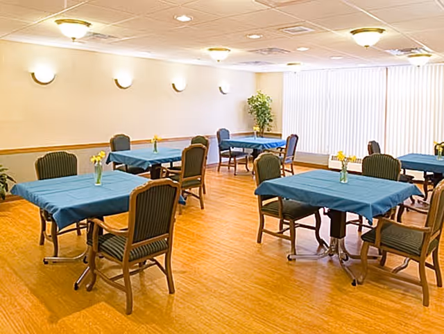 A dining room with several tables covered in blue tablecloths, each table surrounded by chairs with green upholstery. Small vases with yellow flowers are placed on each table. The room has wooden flooring, cream-colored walls, ceiling lights, and large windows with vertical blinds allowing natural light to enter.