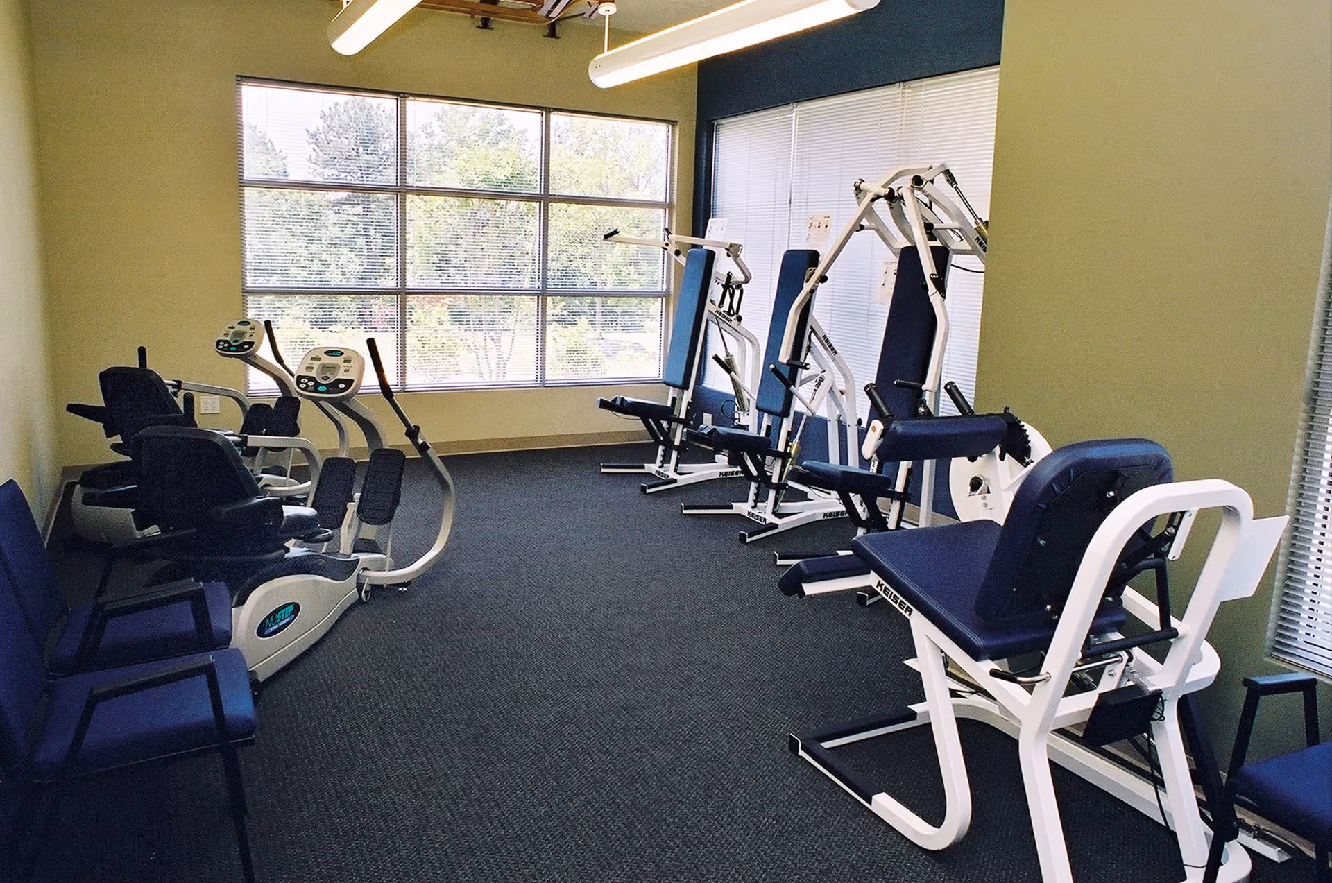 A small fitness room with exercise machines including recumbent bikes and strength training equipment. The room has large windows with blinds letting in natural light and blue cushioned chairs along the walls.
