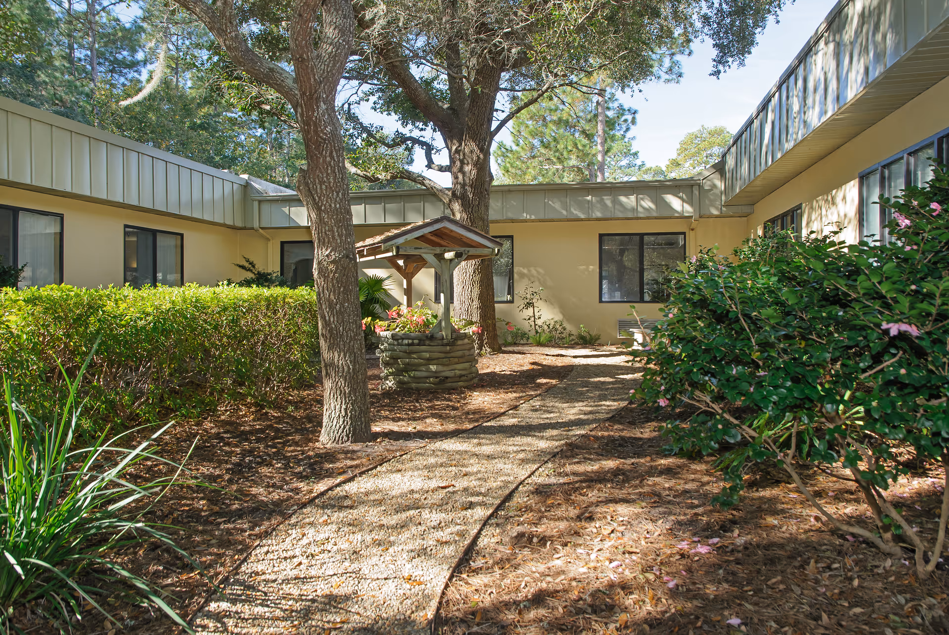 Gravel pathway through a shaded courtyard with trees, shrubs, and a small wooden well between single-story building wings.