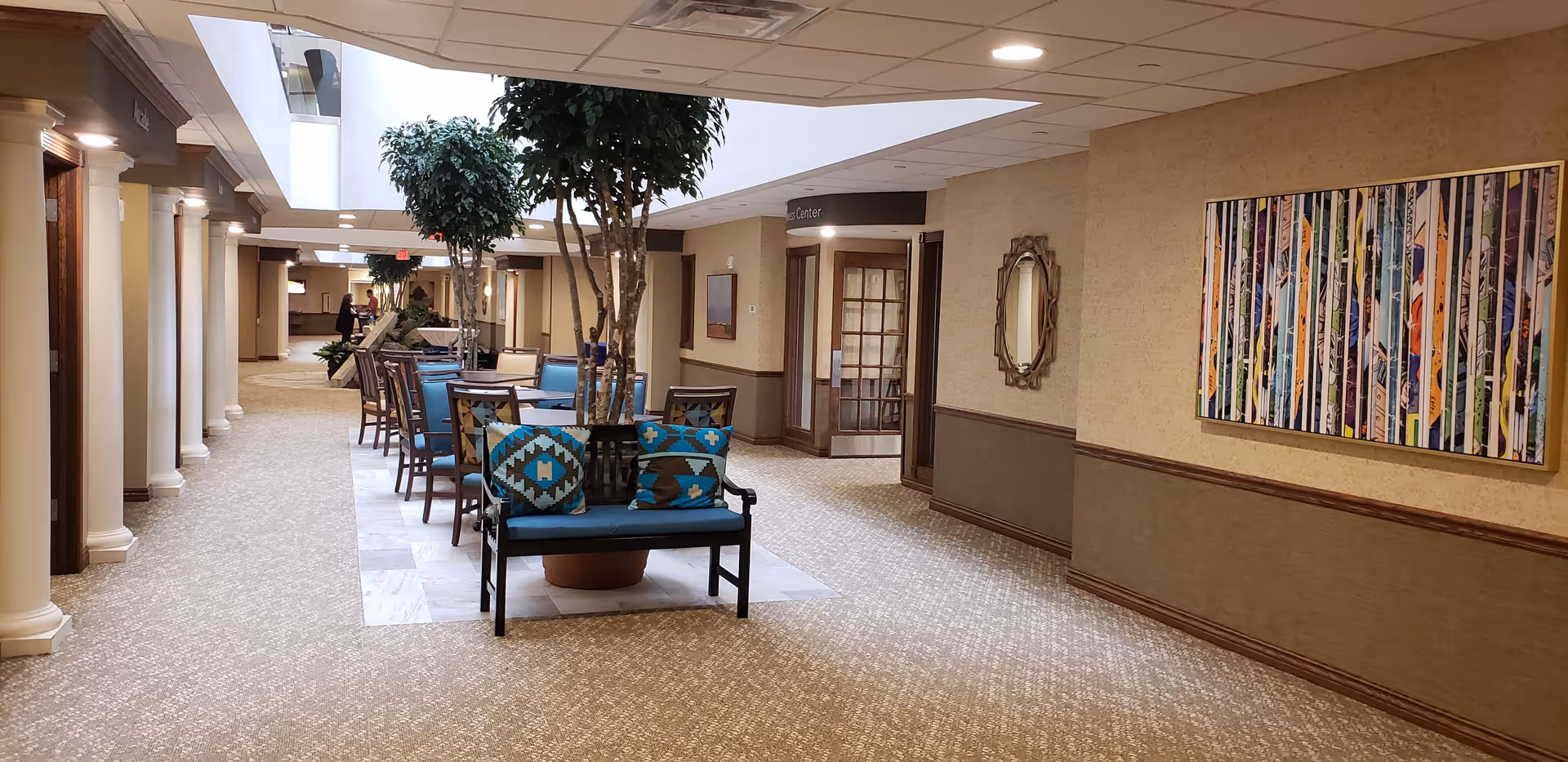 Interior hallway of Daniel Pointe Retirement Community featuring a seating area with blue cushioned chairs and a bench with patterned pillows around a large potted tree. The hallway has beige walls with a decorative mirror and colorful artwork, carpeted floor, and ceiling lights.
