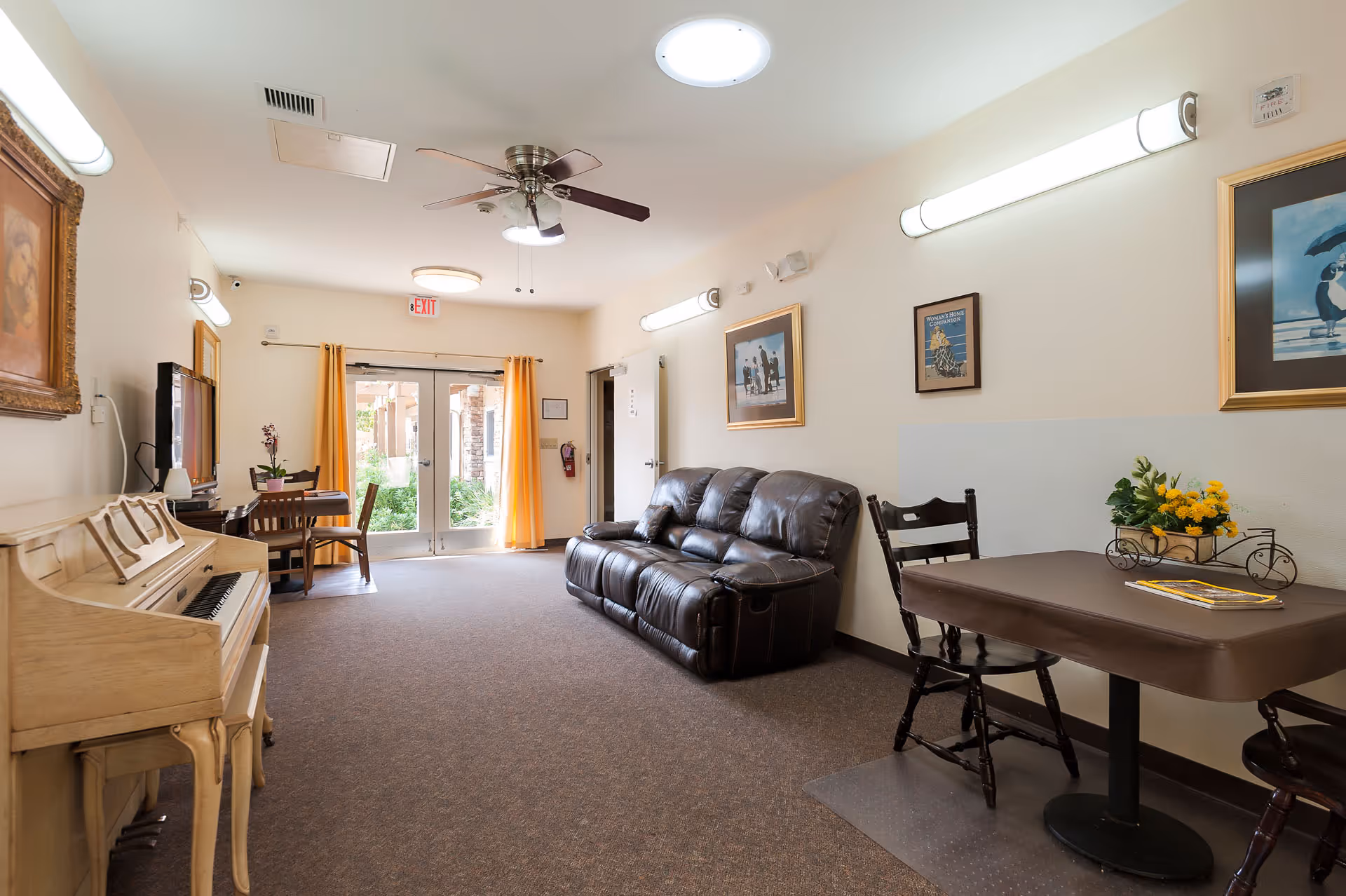 Community lounge with a piano, leather sofa, tables and chairs facing glass double doors to an outdoor area.