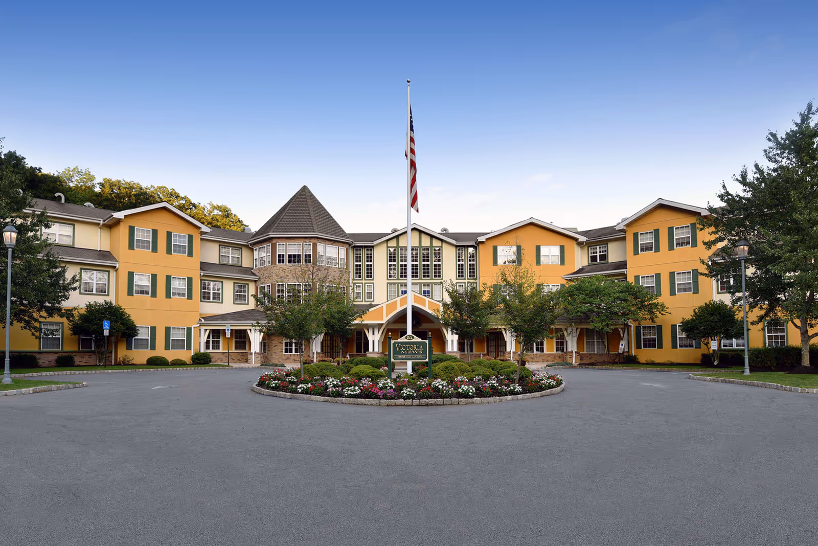 Front exterior view of Victoria Mews Assisted Living facility with a circular driveway, landscaped flower bed, American flag on a flagpole, and a three-story building with yellow and beige walls, green window shutters, and a turret-like structure in the center.