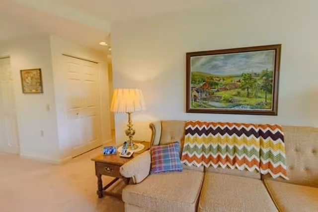A cozy living room area featuring a beige upholstered sofa with a colorful chevron-patterned throw blanket and a plaid pillow. Next to the sofa is a wooden side table with a brass lamp, a phone, and a small framed photo. A framed landscape painting hangs on the wall above the sofa. The room has light-colored walls and carpet, with a hallway visible in the background.