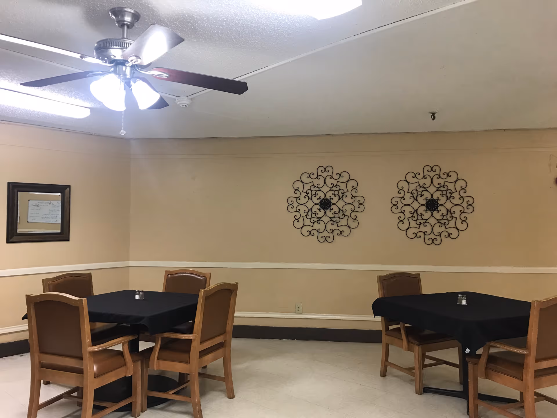 A dining area with two square tables covered with black tablecloths, each surrounded by four wooden chairs with brown cushions. The walls are beige with a white chair rail, decorated with two black ornamental wall hangings and a framed mirror. A ceiling fan with lights is mounted on the ceiling.
