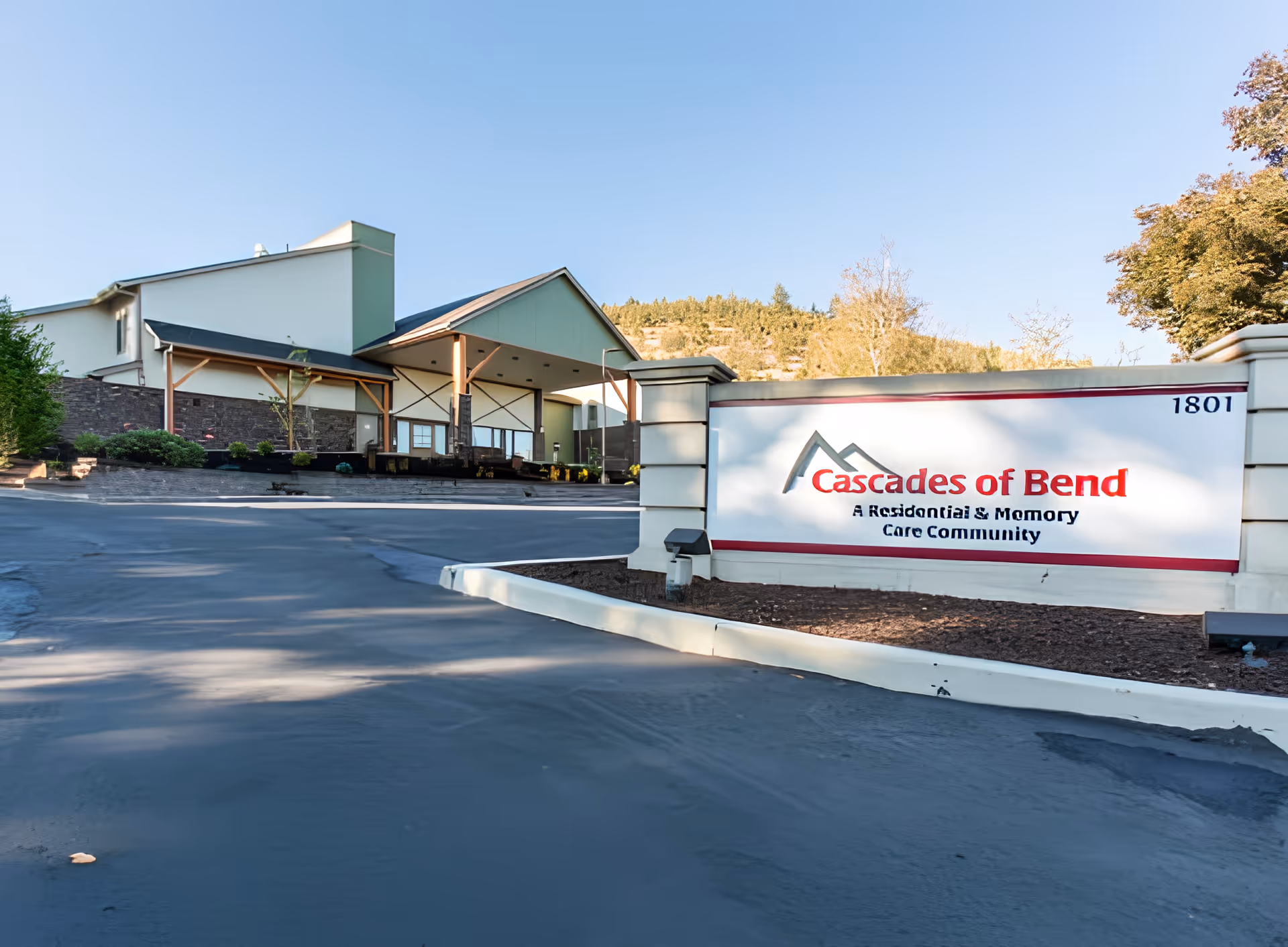 Exterior view of Cascades of Bend Retirement Community building with a clear blue sky and surrounding trees. A large sign in the foreground reads 'Cascades of Bend A Residential & Memory Care Community' with the address number 1801.