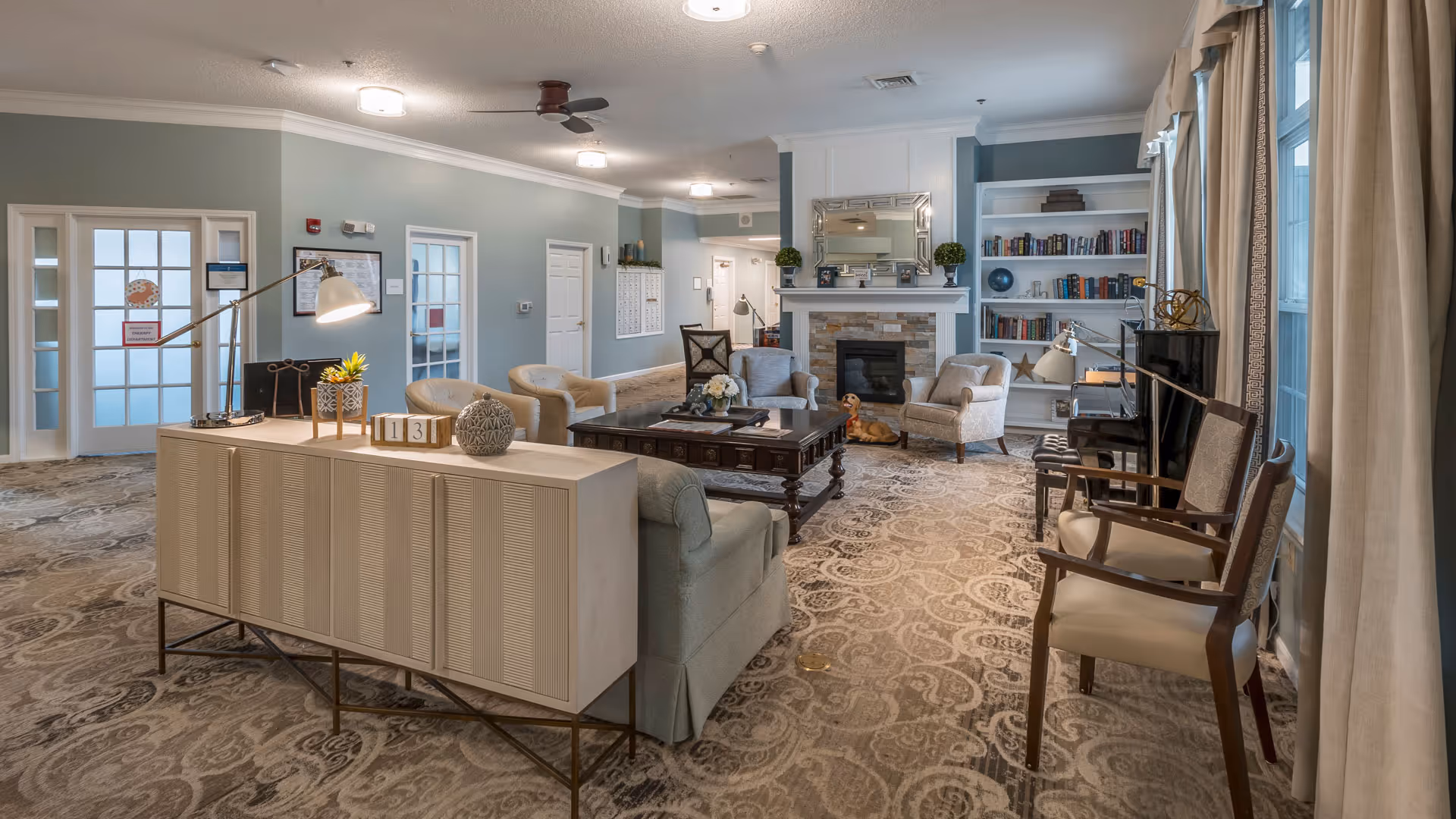 A spacious and well-lit living room area in a senior living facility featuring comfortable armchairs and sofas arranged around a dark wooden coffee table. There is a fireplace with a decorative mirror above it, flanked by built-in bookshelves filled with books and decorative items. The room has patterned carpeting, soft blue-gray walls, and large windows with curtains allowing natural light to enter. A small dog is lying on the floor near the fireplace. Various lamps and decorative plants add to the cozy ambiance.