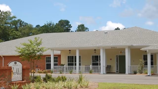 Exterior view of BeeHive Homes of Lynn Haven, a single-story senior living facility with a covered porch, white railings, and a brick and beige facade. There is a sign in front of the building with the facility's name and landscaping with small plants and a tree.