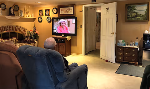An elderly person sitting in a blue recliner chair watching television in a cozy living room with beige walls, decorated with framed pictures, a fireplace, and a wooden cabinet with various items on top.
